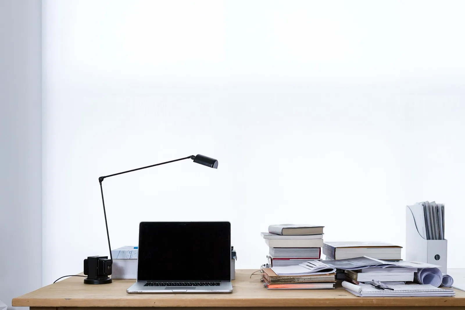 Mesa clara com um laptop, uma luminária e pilhas de livros